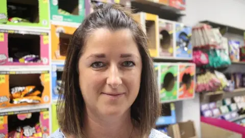A woman with a brown bob and a topknot smiles at the camera, behind her is a wall of orange, green, pink and blue boxes which dispense crisp packets to customers, and packets of sweets.