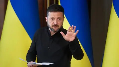 Ukraine's leader wearing black stands in front of Ukrainian blue and yellow flags