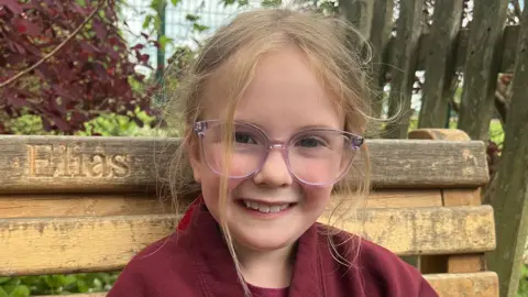 Lola, a young girl with glasses is smiling whilst sitting on a bench with her brother's name carved into the wood. 
