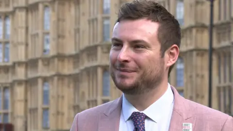 Peter Swallow stands outside the Houses of Parliament, which are out of focus in the background behind him. He looks slightly off camera, smiling, has short spiked light brown hair and a short beard. He is wearing a pink linen suit with a white shirt and navy blue and pint patterned tie.