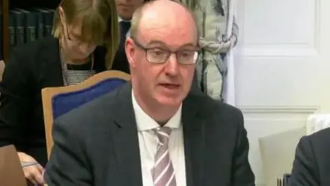Neil Guckian is wearing a pink and white striped tie and wearing black-rimmed glasses. He is wearing a black jacket and is sitting in a purple and brown chair in a room with a number of people.