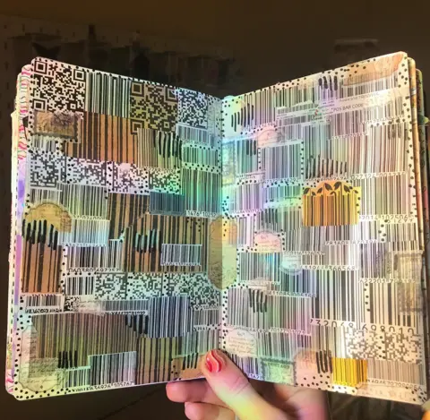 Kia Hunt Two pages of a book are being held open with a hand. They are iridescent barcodes and QR codes covering the whole page.