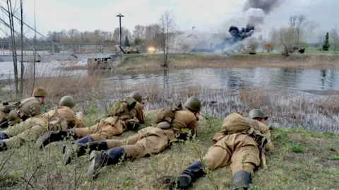 BBC A photo showing men wearing World War Two military uniforms lying on their stomachs in the grass in front of a river. On the other side of the river plumes of smoke are erupting into the cloudy sky