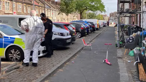 Three police officers are standing around the bonnet of a police car, which is parked in a line of other cars. Behind them is a footpath with various items on it.