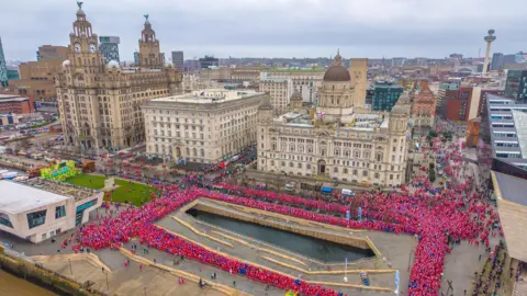 Stratus Imagery Aerial drone view of thousands of participants in red Santa suits on the Liverpool Pier Head with the Three Graces buildings and city skyline behind. They form a rectangular shape around a canal lock near the Mersey waterfront.