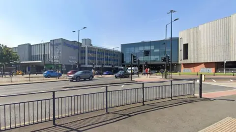 A road with several lanes. Shopping centre buildings on the far side of the road. Railings on the near side of the road. People can be seen on a crossing island in the middle of the carriageways.
