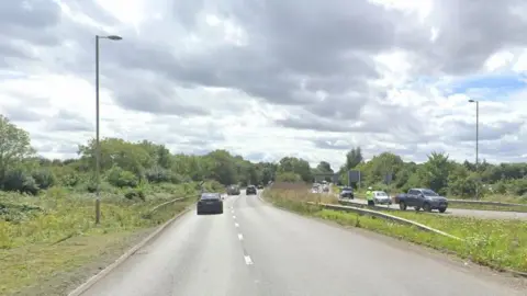 A Google street view of the A41 Oxford Road looking in the direction of the M40 roundabout (not visible). It is a standard looking dual carriageway and has trees on either side.