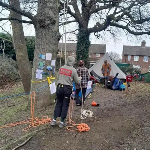 Gabi Barrett People standing at the camp - one person is holding orange rope ready to climb the tree. There are posters and poems on the tree, camp gear on the floor and a bell tent in the background and behind that there are houses which are part of the housing estate