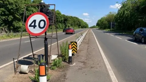 The central reservation of a dual carriageway with a 40 sign in a red circle on a metal stand. There are portable light sticks in black, white and red on the ground, and a low metal barrier stretching beyond. There are trees either side of the road and two cars are heading away from the camere on the left.