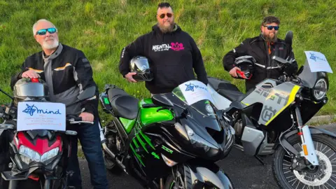 Christopher Smith is pictured with his father and brother in law. All three are wearing biker gear and holding their helmets under their arms. They are smiling at the camera while standing next to their motorbikes. Each motorbike has a small sign on the front that says Mind with a little blurb about their fundraiser.