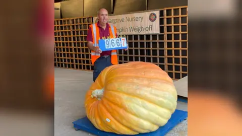 Curtis Leach Curtis Leach standing up this time, next to a giant pumpkin with a sign saying "966.7", which signifies that it weighed 966.7lbs