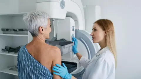 Getty Images A female worker in a white lab coat and blue gloves screens a lady with short grey hair. The worker has her left arm touching the right arm of the patient. They are looking at each other. The patient has a hospital gown covering half of her upper body. She is standing at the white machine to the front of her. There are white shelves to the left