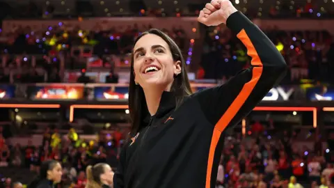 Caitlin Clark photographed gesturing towards the crowd from the court during WNBA All-Star weekend. She is smiling and wearing a black sports top trimmed with orange. 