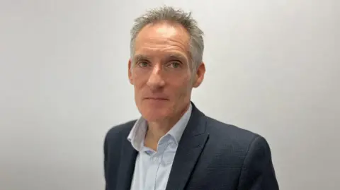 The photo shows Nick Hynes looking at the camera with a blank wall background. He has short greying hair and is wearing a dark suit and a pale shirt.