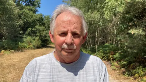 BBC/David Allard Ian Knight with short grey hair wearing a light grey Adidas T-shirt, standing on a sunlit woodland path. Trees and green foliage line both sides of the trail under a clear blue sky.