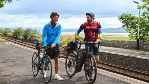 Two cyclists smiling as they push their bikes down a  section of the route near Kent's Bank Station in Grange-over-Sands. The rail line is on their right, with the coast visible in the distance.