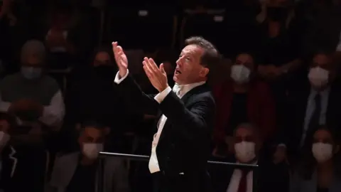 Welwyn Garden City Music Society James Ross in formal concert attire stands at a podium with raised hands, directing a performance. In the background, an audience sits in dim lighting, many wearing face masks, watching the event.