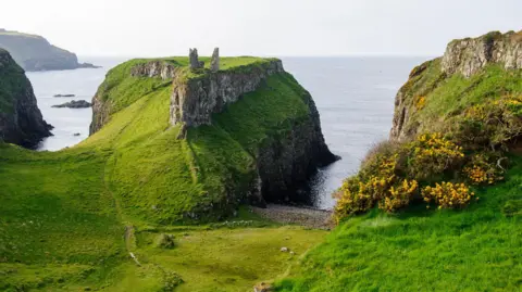 Getty Images Dunseverick Castle sits atop a headline on the north coast, which is grassy and rocky. The remains of the castle are visible and are two stone walls on either side.