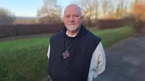 Father Jan Rossey stands successful  a accepted   monk's wont  - achromatic  robe covered successful  a achromatic  hooded robe with a transverse  connected  a agelong  concatenation  astir   his neck. His hands are down  his backmost  and he's looking into camera, smiling slightly. He is bald with a achromatic  goatie beard.