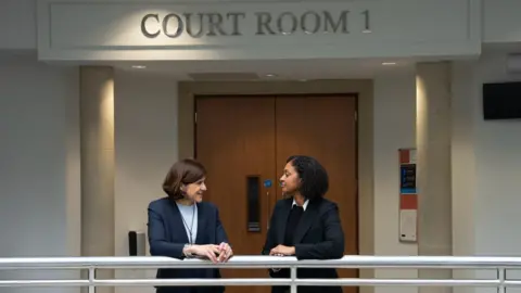 Two women in black suit jackets talk on a balcony with courtroom signage above their heads. Brown double doors can be seen in the background. 