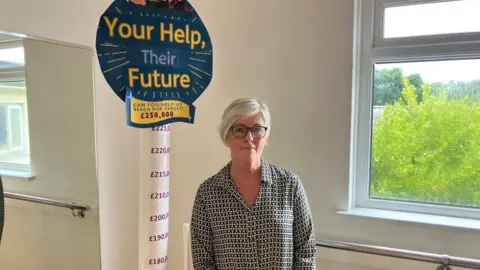 A woman stands in an old dance studio in front of a fundraising totalizer . She wears a grey shirt and glasses