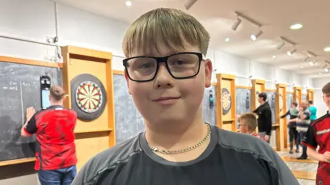 Dylan is smiling - he has short brown hair and is wearing square framed glasses and a black and grey top and a silver chain. Behind him is a hall lined with darts boards with other young people throwing darts. 