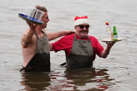 PA Media Two men in aprons stand waist-deep in the water. They both wear aprons and carry trays with drinks. One also wears a red t-shirt and a Santa hat.