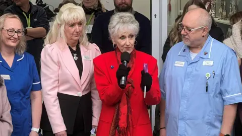 BBC Anita Dobson looking at the camera wearing a red coat and scarf with black gloves. To her left is Julie Rowley, Director of retail at Thames Hospice she is wearing a black dress and pink short jacket.