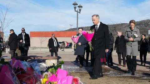 Getty Images An image showing Carney, who is dressed in a black coat and black trousers, holding a bouquet of flowers wrapped in pink paper. He is walking towards a pile of flowers at a memorial for victims of the mass shooting in Tumbler Ridge. Behind him is his wife, Diana Carney, also holding a smaller bouquet. 
