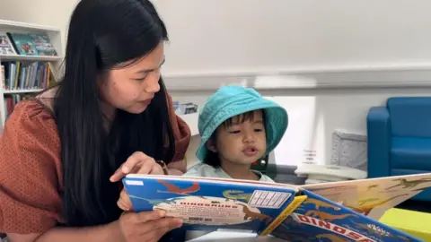 A mother and her daughter read a book. The mother has long black hair, and is wearing a brown/rust coloured top. She is holding a book that her young daughter appears to be reading. The child has shoulder length dark hair, and is wearing a white top and a turquoise sun hat. There is a book in the foreground, with the word "dinosaur" visible. A blue sofa is to the centre right of the image, and book shelves to the left.