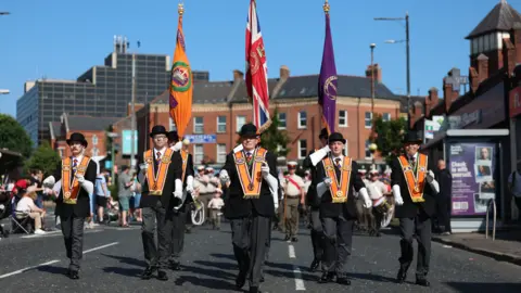PA Media Five men wearing Orange Order sashes are leading a march the middle of a road. Three behind them are holding flags. Crowds are in the background. 