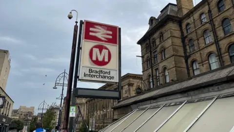 Bradford Interchange, with a view looking up to a sign indicating it is both a bus and a rail station. In the distance is St George's Hall and other city centre buildings. 