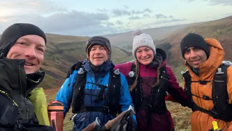 Runners Chris Parr, Paul Baxter, Eva Kriki and Behzaad Ghouse stand together a top of fells in the north. They are wearing trial running equipment and clothing of various bright colours.