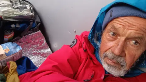 Selfie of Nick Brooke wearing a red coat and another blue waterproof jacket underneath with the hood up. He appears to be laying on the floor and leaning on a wall, surrounded by his belongings, including a rucksack. He has a tanned face and a full grey beard.