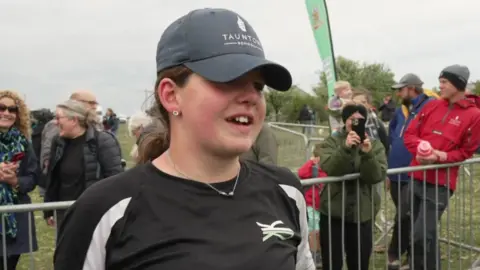 Josie is wearing a cap that has TAUNTON SCHOOL on it. She is being interviewed. Behind her are adults watching her from behind a metal railing.