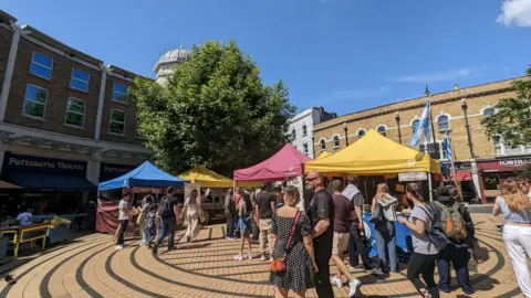 A row of multicoloured stalls surrounded by people in a pleasant looking plaza.