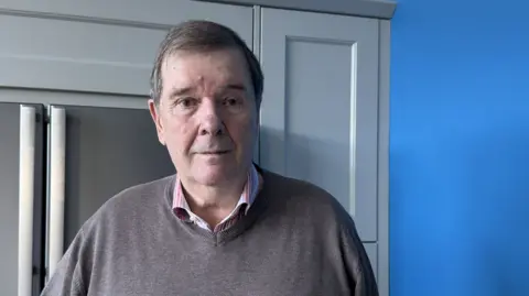 David Czarnetzki at home, wearing a shirt and brown jumper. He is looking at the camera with a worried expression.