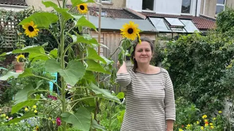 Harriet smiles into the camera, with tall sunflowers behind her, standing in a garden. She is wearing a grey striped dress and her brown hair is tied back.