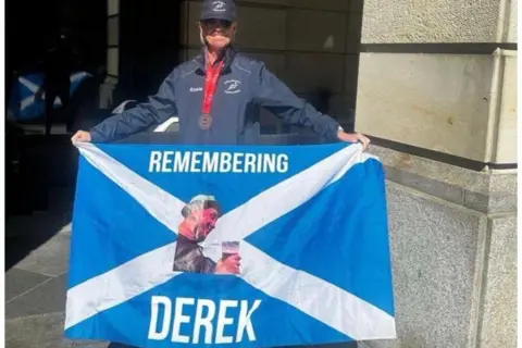 Rosie McGrane Rosie McGrane stands with a medal around her neck holding out a Scotland flag with the words 'REMEMBERING DEREK' on it along with a picture of her late brother Derek.
