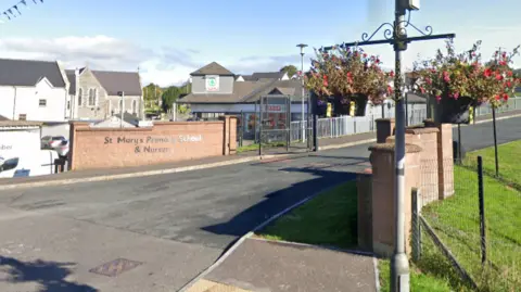 An image of the school gates of St Mary's Primary School. There are two red bricked walls leading into the main entrance. On the red wall a sign reads "St Mary's Primary School & Nursery". A Spar shop can be seen in the background. 