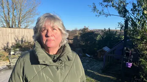 Amy Holmes/BBC A head and shoulders shot of a woman. She is standing in a garden with blue sky behind her. She has grey hair covering her ears and is wearing a light navy green coat. There is part of a tree behind her on the right.