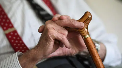 A close-up image showing an elderly man sat down, holding a walking stick. He is also wearing a shirt, tie and braces.