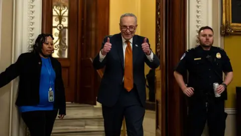 Getty Images Schumer gives a double thumbs up as he walks out of an ornate doorway opened by a female worker on the left and guarded by a male police officer on the right