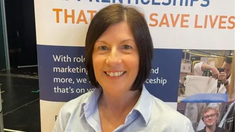 RNLI A smiling Karen Lyall stands in front of an RNLI apprenticeships display board. She has a dark bob haircut and is wearing an open-collar pale blue shirt.
