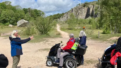 A woman in a black and blue coat is gesturing towards a large rock formation. Looking on are two people, one of whom is riding a mobility scooter