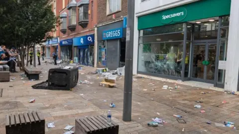 Shops in Hull city centre with smashed windows following rioting in summer 2024. Debris is strewn on the pavement outside.