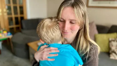 Nikki Fox/BBC Emily is a third year student midwife and is cradling her 6-year-old son in her arms, you can see her head and shoulders and she is looking at him lovingly. Her son has blonde hair and a blue T-shirt. She is wearing a brown jumper and has blonde, long mousey hair which is worn with the sides back. Her fringe is growing out and is parted in the middle. They are in her living room with a sofa and cushions behind her.