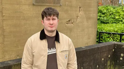 Dan Lonsdale is standing in front of an old stone building and a dark stone wall, with leafy vegetation in the background. He's wearing a cream cotton anorak with brown collar, and a brown Dickies t-shirt. He has short, brown, curly hair and a neutral expression.