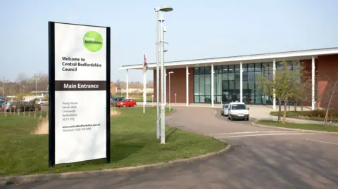 A modern council building sits beside a curved driveway, with a tall sign in front reading “Welcome to Central Bedfordshire Council” and “Main Entrance.” The area has grass, trees, and a few parked vehicles under a clear sky.