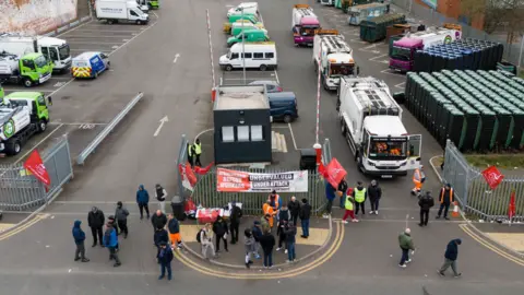 PA Media An aerial view of people standing outside the metal gates of a bin lorry depot. There are signs hung up on the fences and some people are in orange fluorescent clothing. On the right hand side there are two white bin lorries waiting to leave the depot, but people are blocking their way